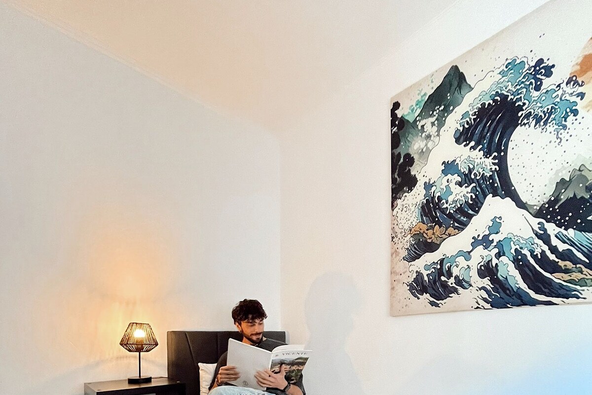 A bedroom with a bed, a nightstand, a lamp, a blanket, and a painting on the wall.