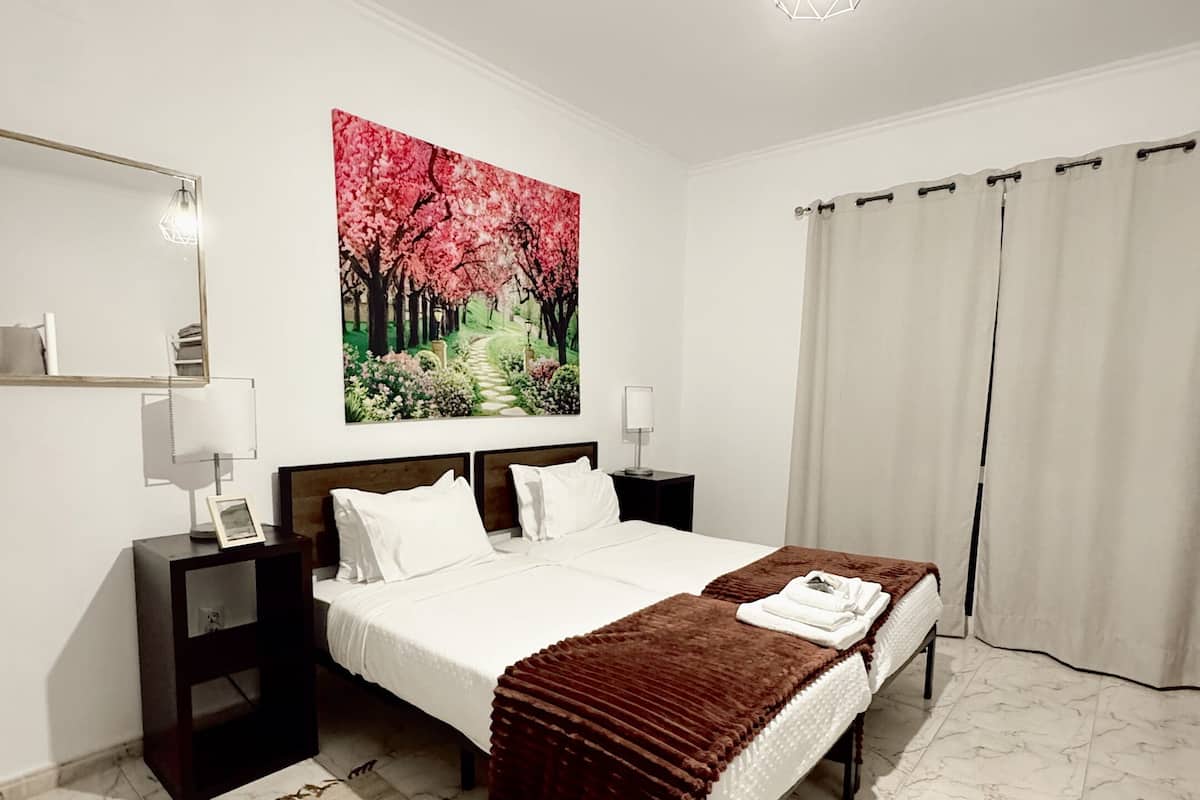 Ein Hotelzimmer mit einem großen Bett, einem Nachttisch, einer Wandlampe, einem Gemälde von einer Kirschblüte und einem Fenster mit Vorhängen.