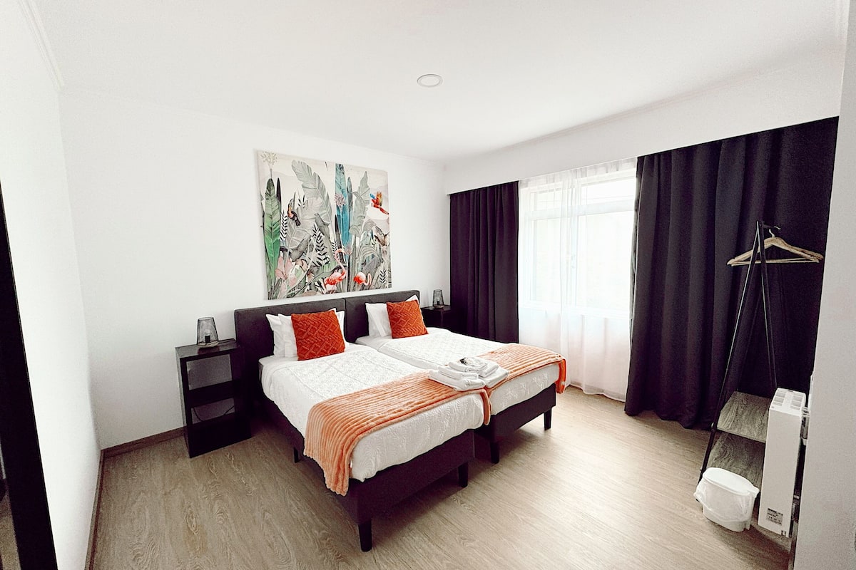 Ein modernes Hotelzimmer mit einem großen Bett, einem Nachttisch, einem Schreibtisch, einem Sessel, einem Kleiderschrank, einem Fenster mit Vorhängen und einem Bild an der Wand.