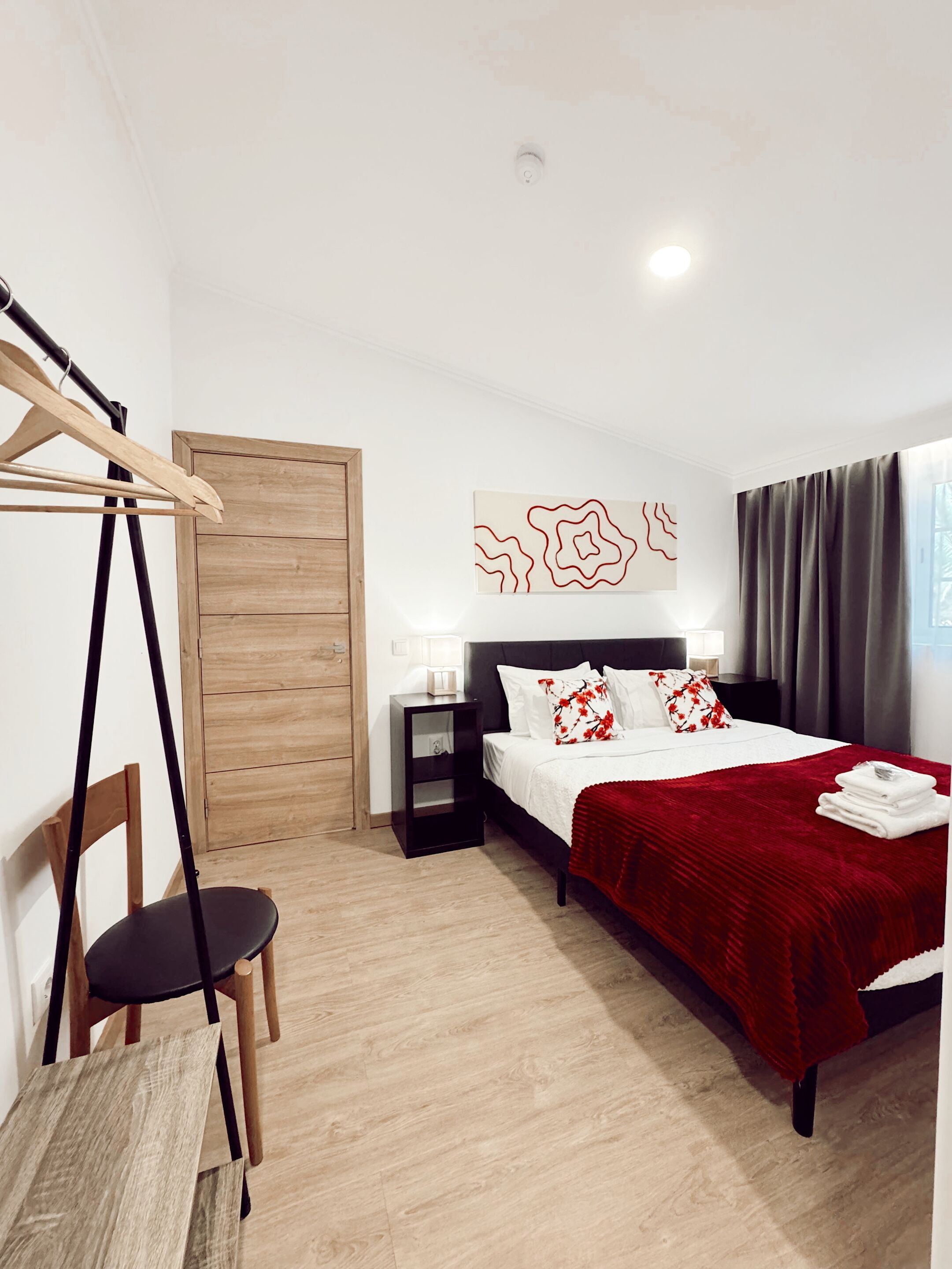 A bedroom with a wooden wardrobe, a bed with a red blanket, a nightstand, a chair, and a wall art piece.