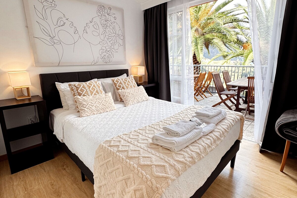 A bedroom with a large bed, a nightstand, a wall art piece, and a view of the outdoors.