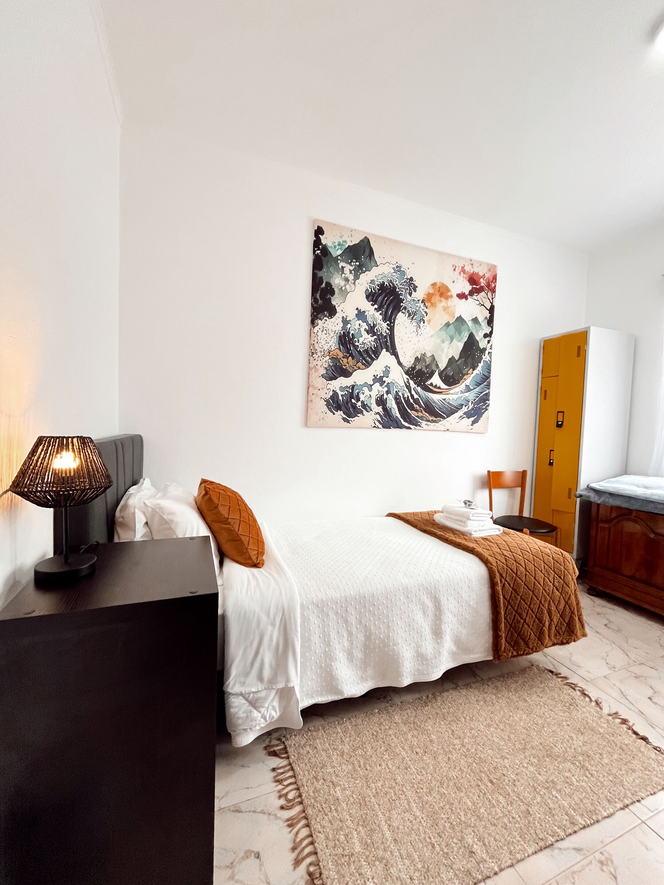 A modern bedroom with a bed, a bedside table with a lamp, a wooden chair, a wardrobe, and a large abstract painting on the wall.