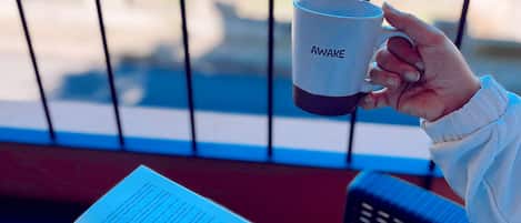 Eine Person hält eine Tasse, auf der das Wort „AWAKE“ steht, und sitzt auf einem Balkon mit Blick auf Gebäude und Grünflächen.
