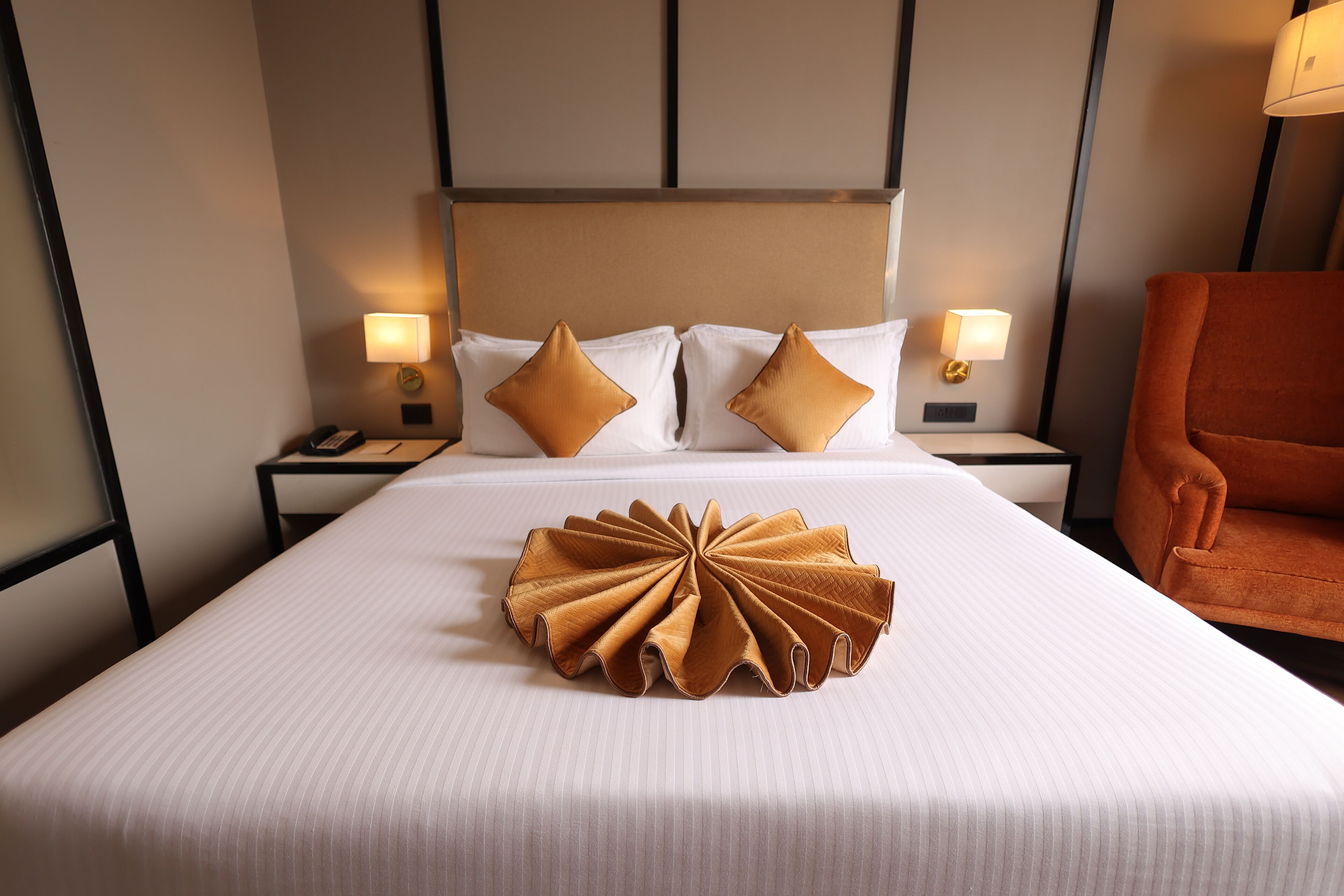 Una habitación de hotel con una cama bien hecha, dos lámparas de noche, un teléfono y un sillón naranja suave.