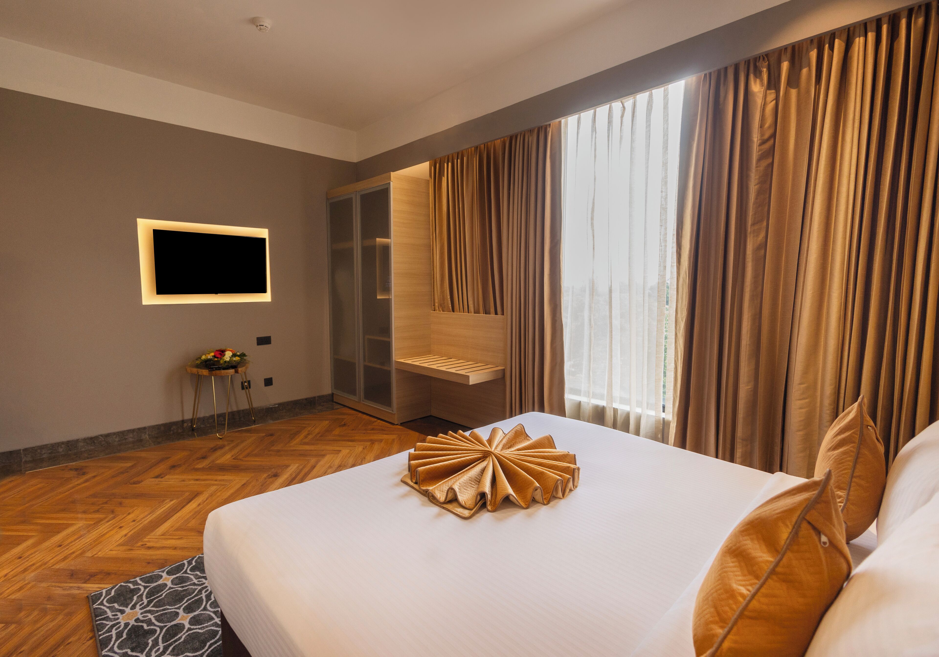 A hotel room with a large bed, a flat-screen TV mounted on the wall, and a view of the city through large windows.