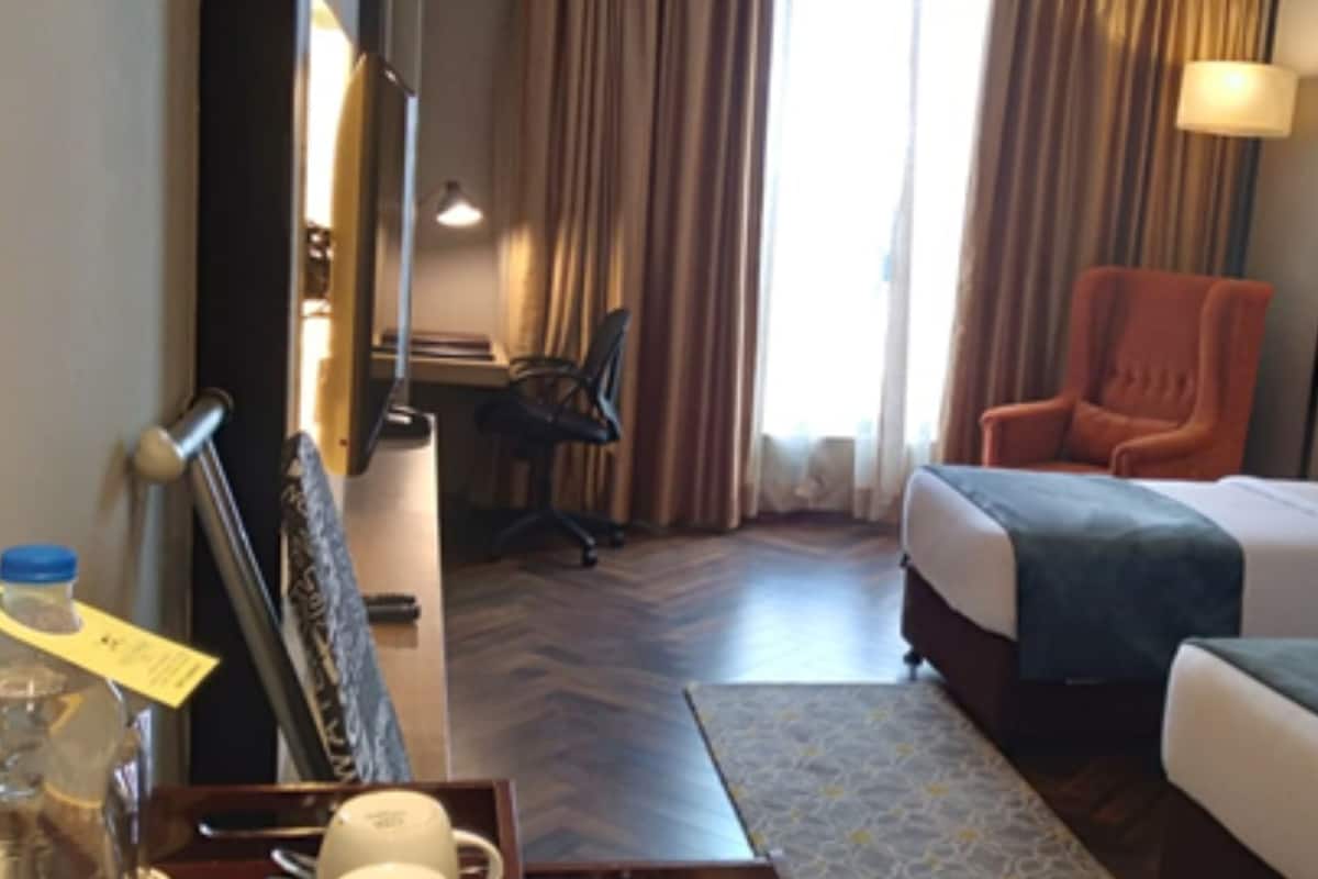 Ein Hotelzimmer mit zwei Betten, einem Schreibtisch, einem Stuhl und einem fensterbepflanzten Fenster.