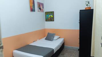 Quarto pequeno com uma cama de solteiro, decoração em branco e laranja, um guarda-roupa preto e duas obras de arte nas paredes.