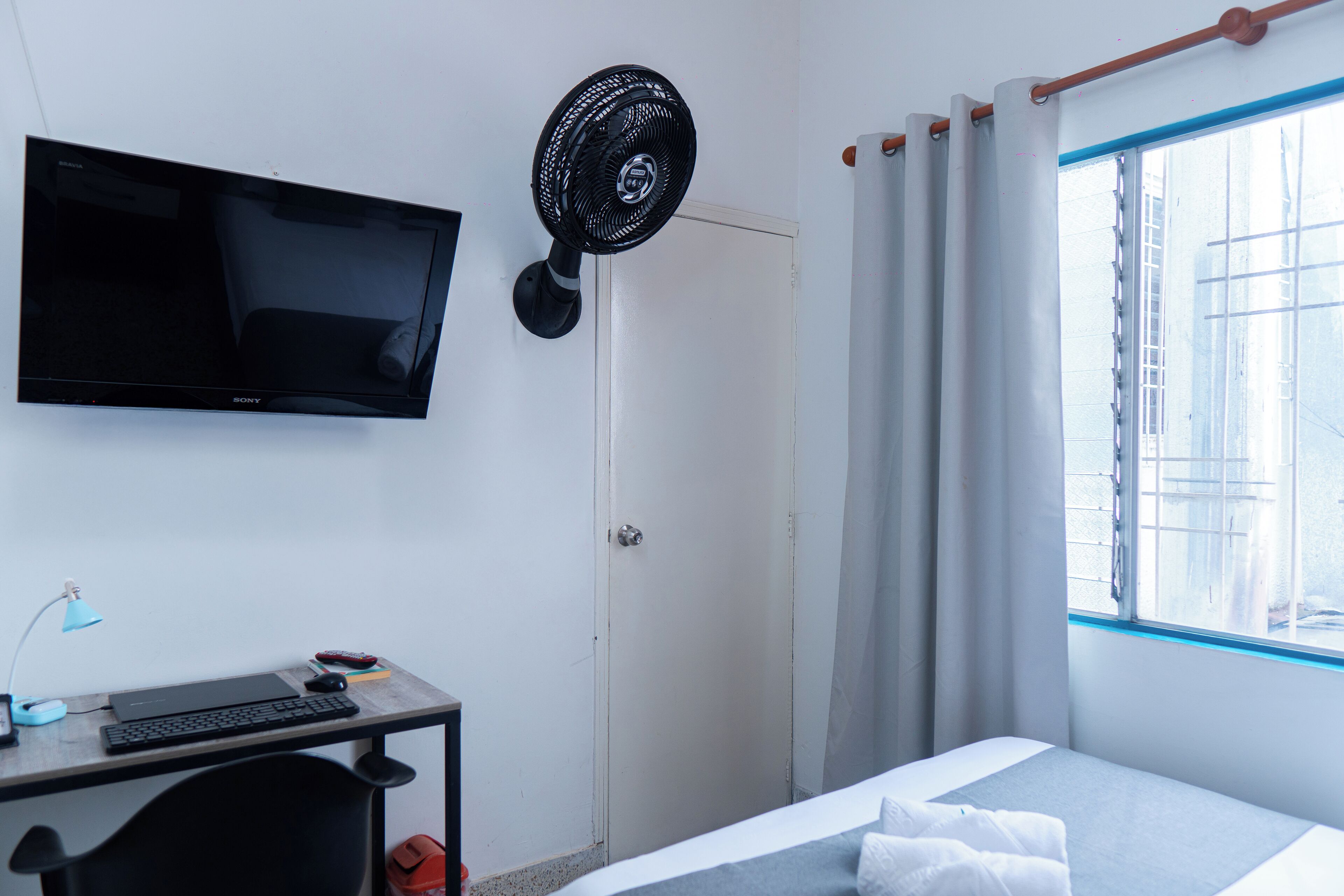 Hotellihuone, jossa on taulutelevisio, lattiatuuletin, työpöytä kannettavalla tietokoneella ja valkoisilla vuodevaatteilla varustettu sänky.