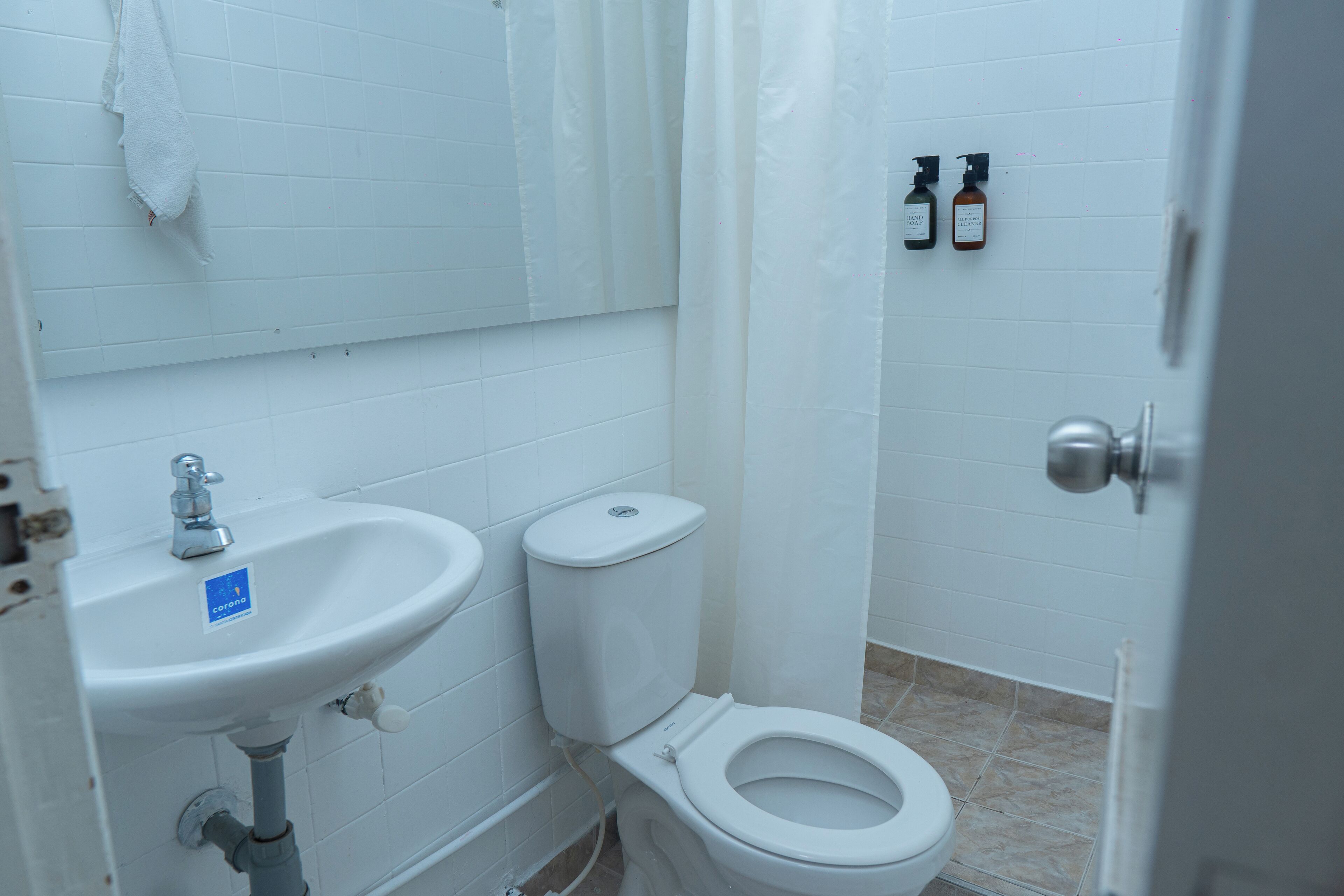 Un baño con lavamanos blanco, inodoro y cortina de ducha. Hay dos botellas en la pared.