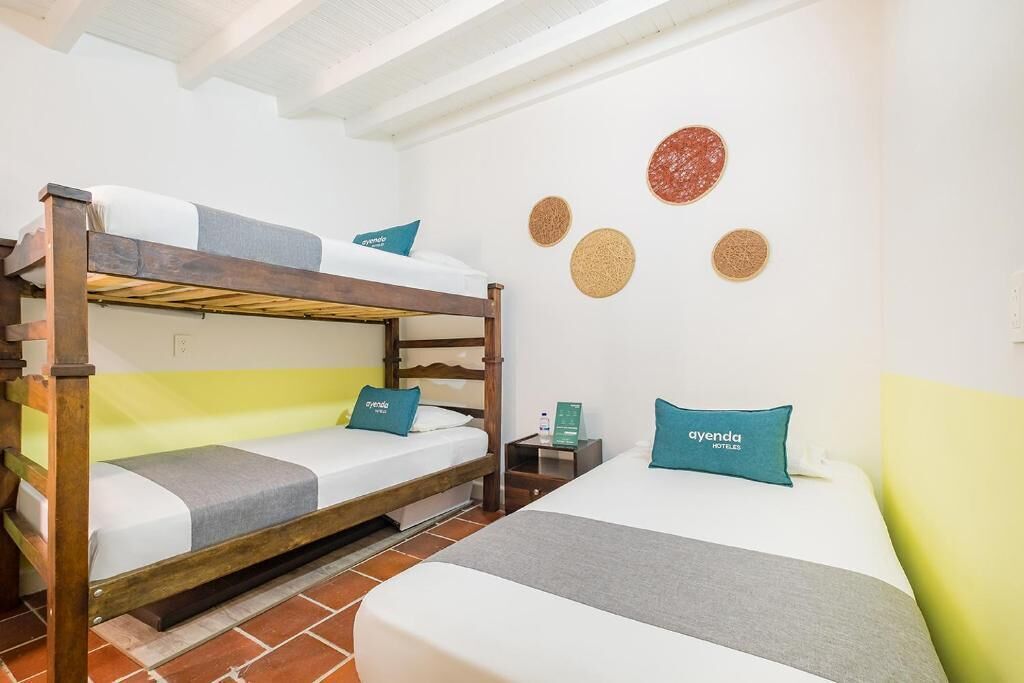 Habitación de hotel con litera, cama individual y mesita de noche con lámpara.