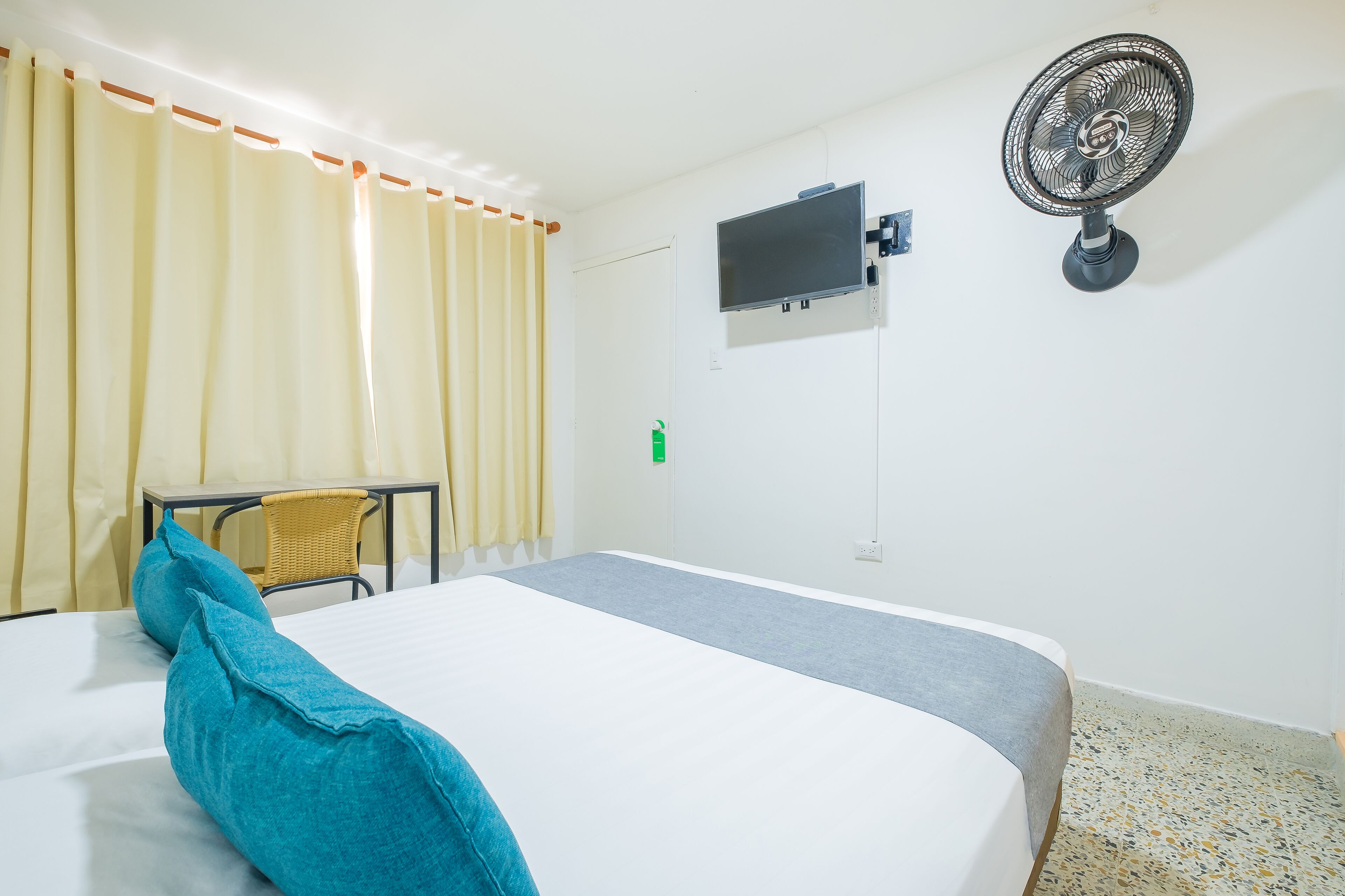 Habitación de hotel con cama, escritorio, silla, televisor y ventilador de pared.