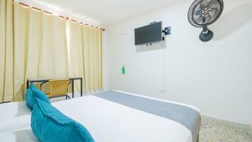 Habitación de hotel con cama, escritorio, silla, televisor y ventilador de pared.