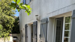 Un patio exterior con mobiliario de mimbre, suelo de piedra y un edificio con contraventanas grises y una puerta.