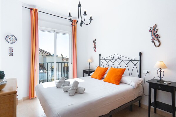 A hotel room with a bed, two bedside lamps, a chandelier, a balcony with curtains, and a view of buildings outside.