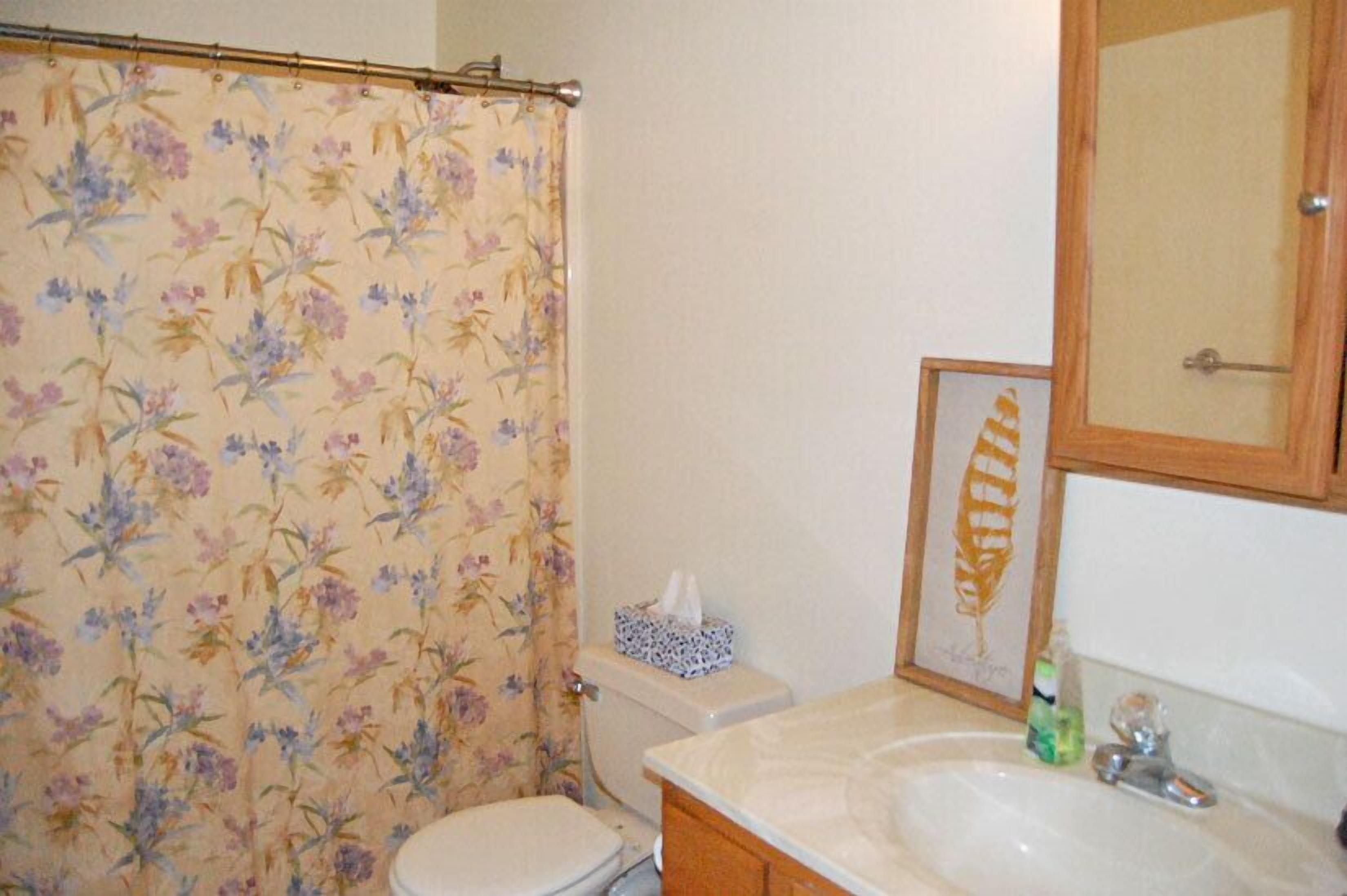 Un baño con cortina de ducha estampada con motivos florales, un inodoro, un lavamanos con espejo y un adorno con una hoja enmarcada.