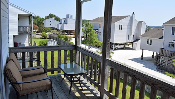 Een houten terras met een glazen tafel en twee stoelen, met uitzicht op een woongebied met huizen en een heldere lucht.
