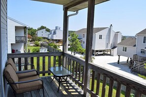 Um deck de madeira com mesa de vidro e duas cadeiras, com vista para uma área residencial com casas e céu claro.