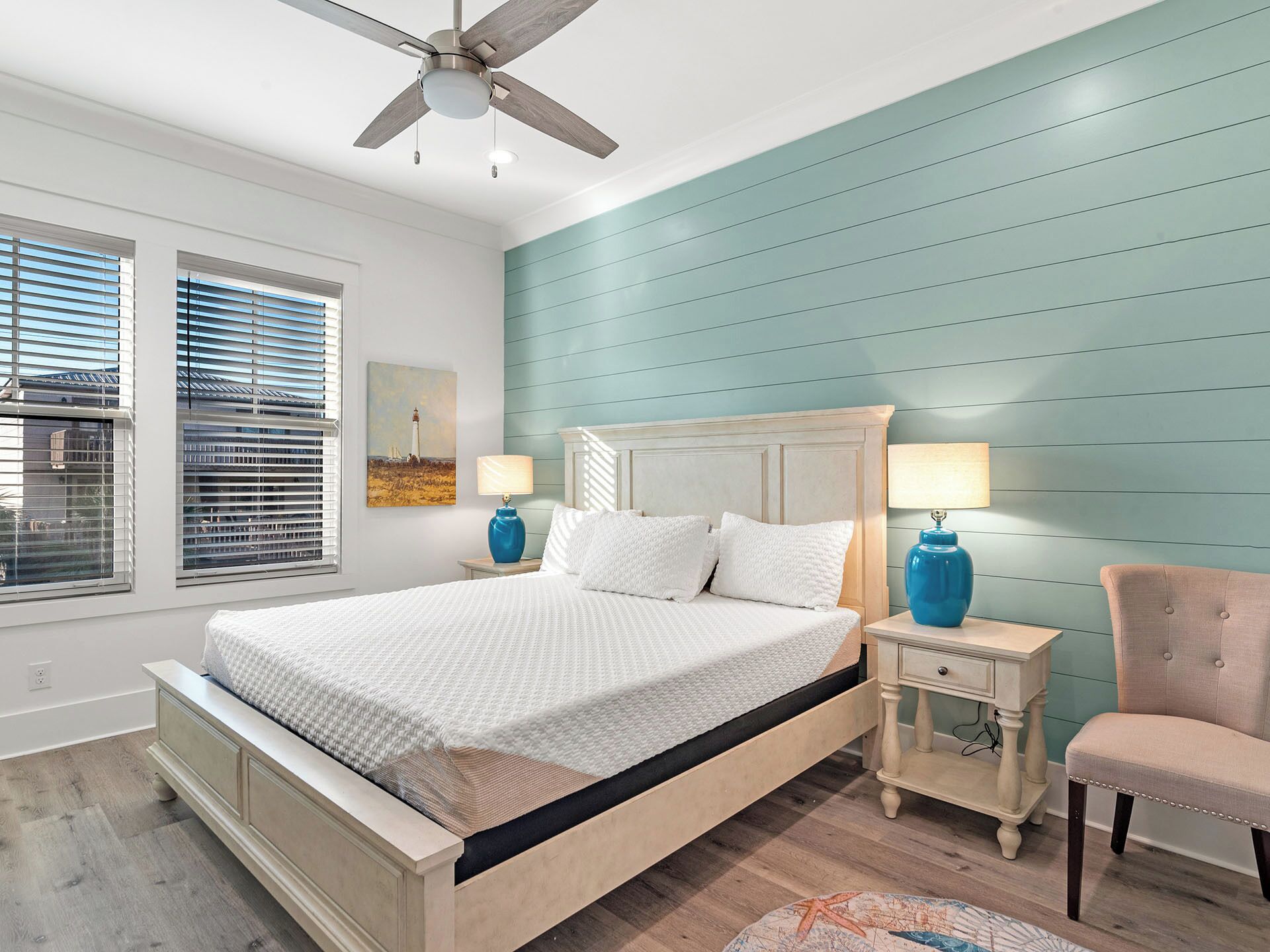 A bedroom with a bed, two bedside tables with lamps, a chair, a ceiling fan, and a window with blinds.