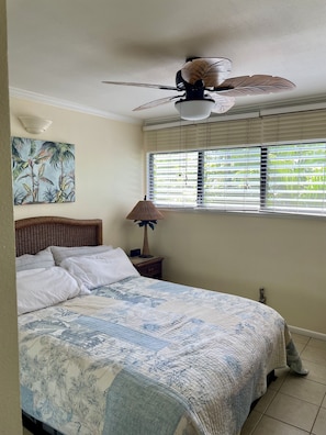 A bedroom with a bed, a ceiling fan, a window with blinds, a wall-mounted lamp, and a painting.