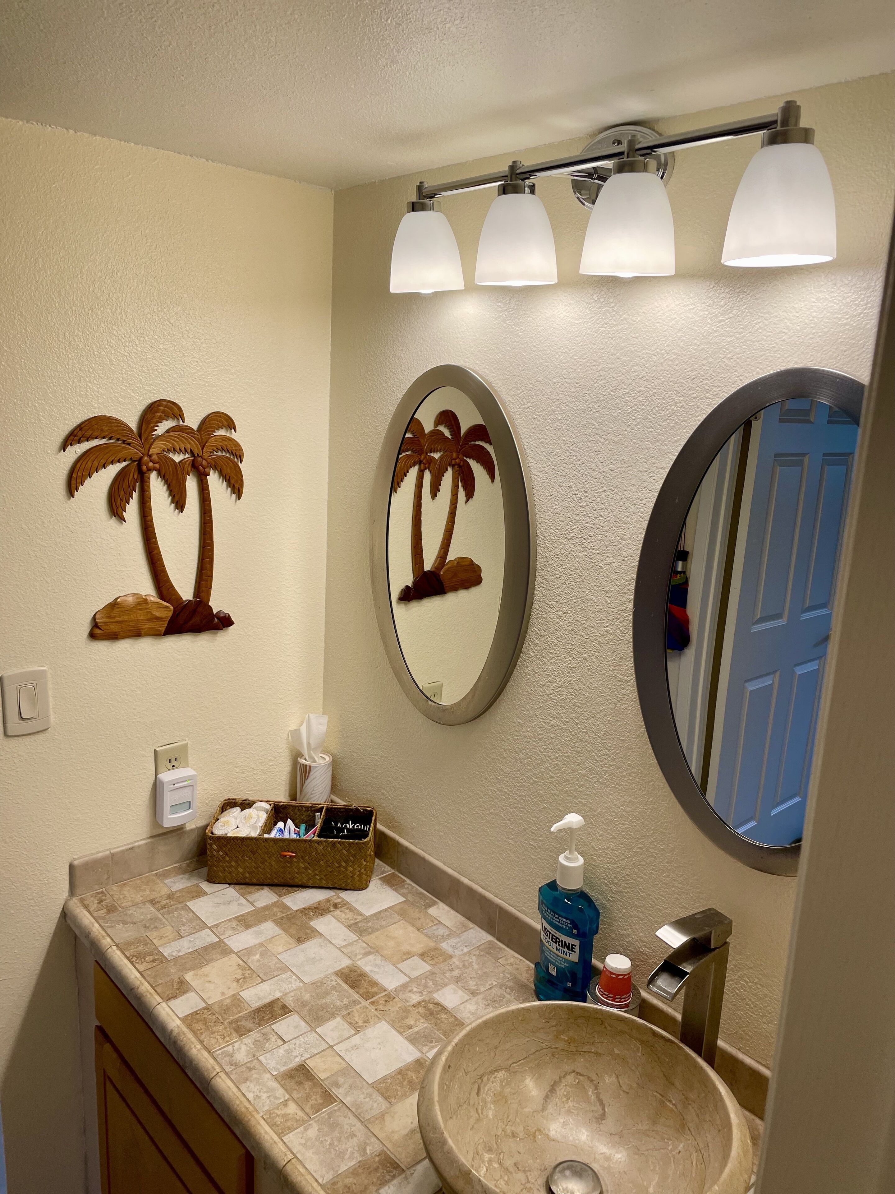 A bathroom with a large oval mirror, a wall-mounted light fixture, a sink with a beige basin, and a decorative wall piece of palm trees.