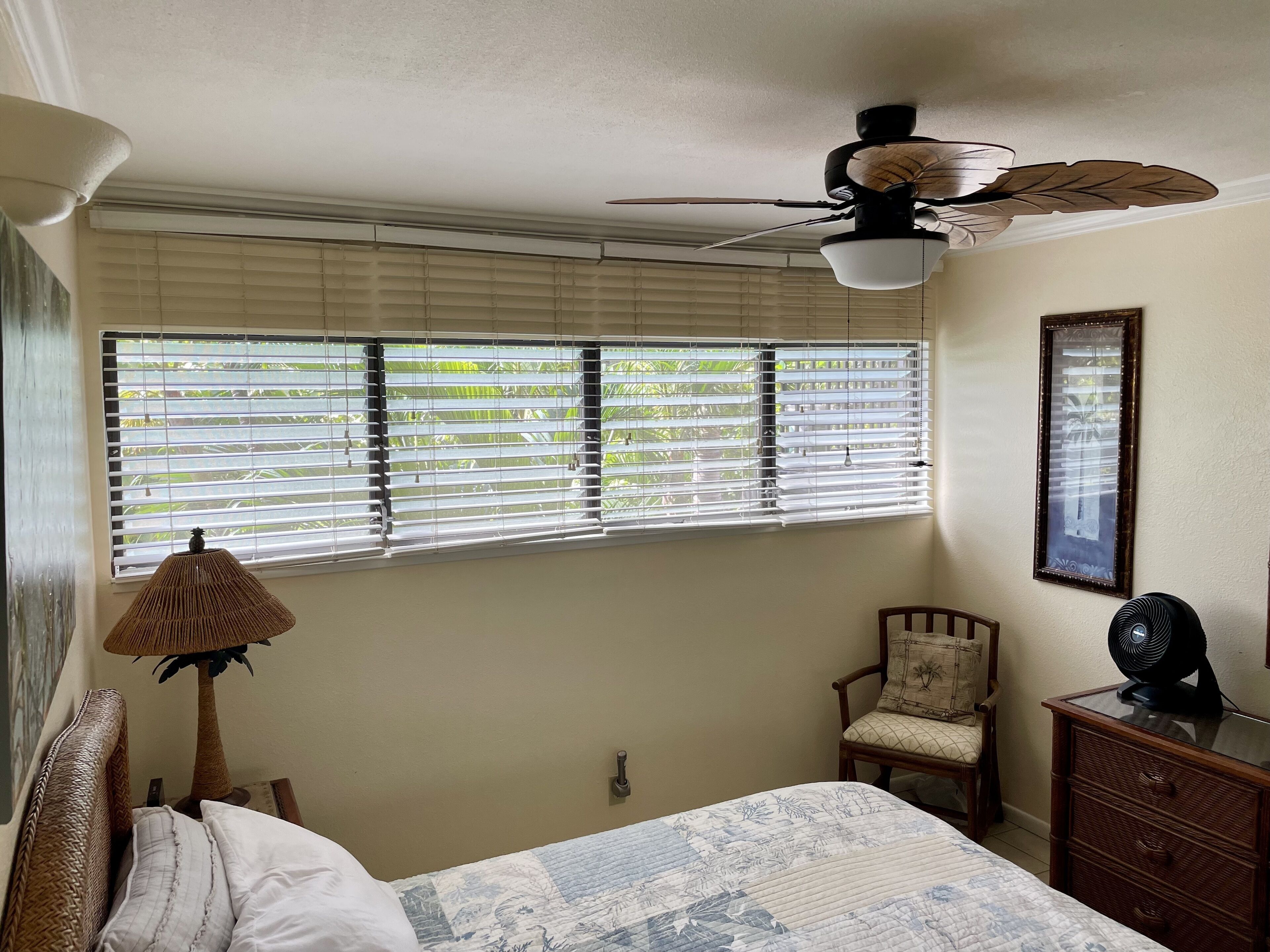 Une chambre à coucher comprenant un lit, une chaise, une commode, un ventilateur de plafond et une fenêtre avec des stores.