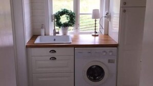 Pieni ja käytännöllinen pyykinhuoltotila, jossa on pyykinpesukone, pesuallas ja ikkuna, jonka ympärillä on koristeellinen seppele.