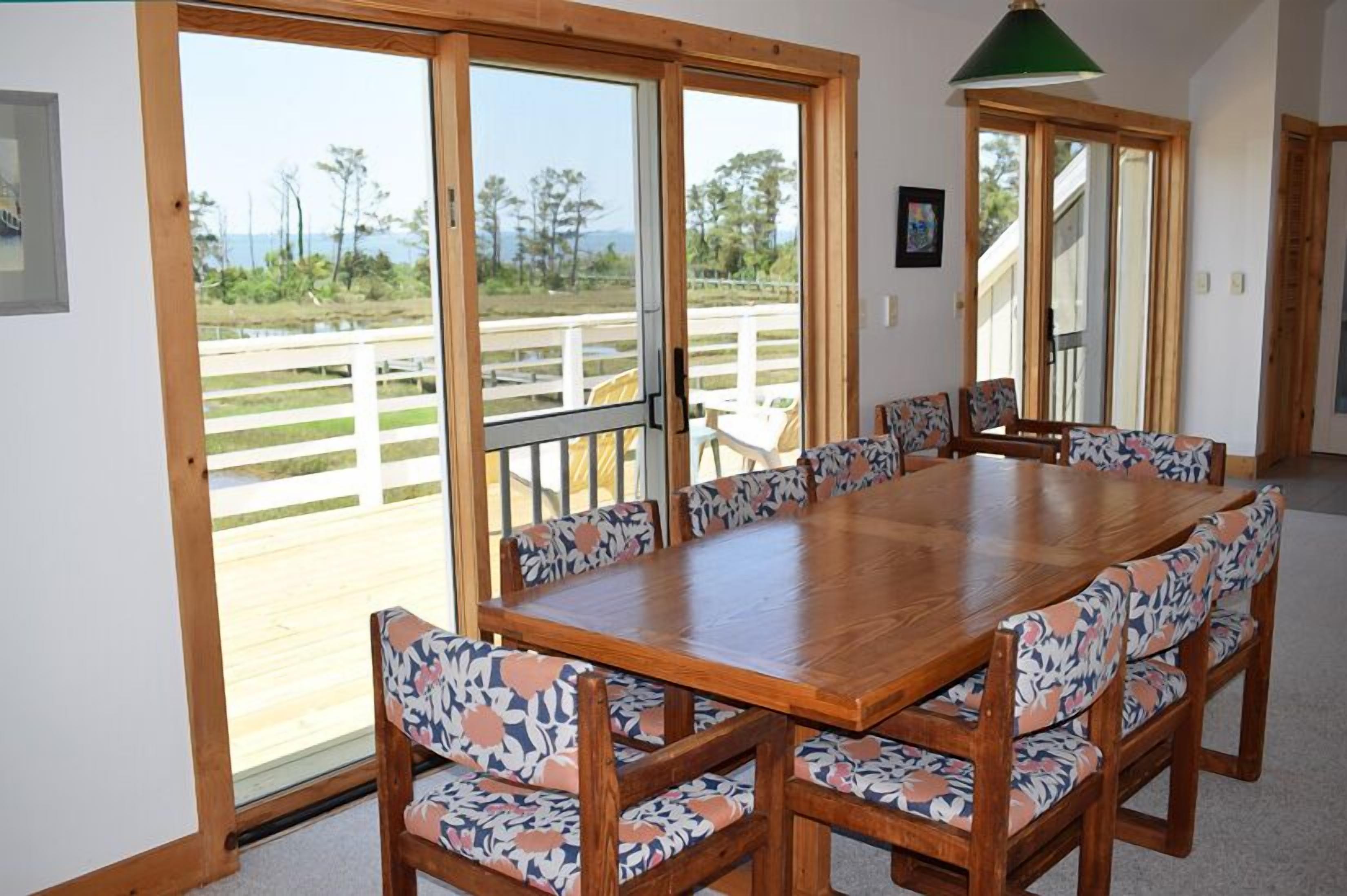 Un coin repas avec une table et des chaises en bois, de grandes portes vitrées donnant sur une terrasse agrémentée d’une clôture blanche, et un luminaire suspendu vert.