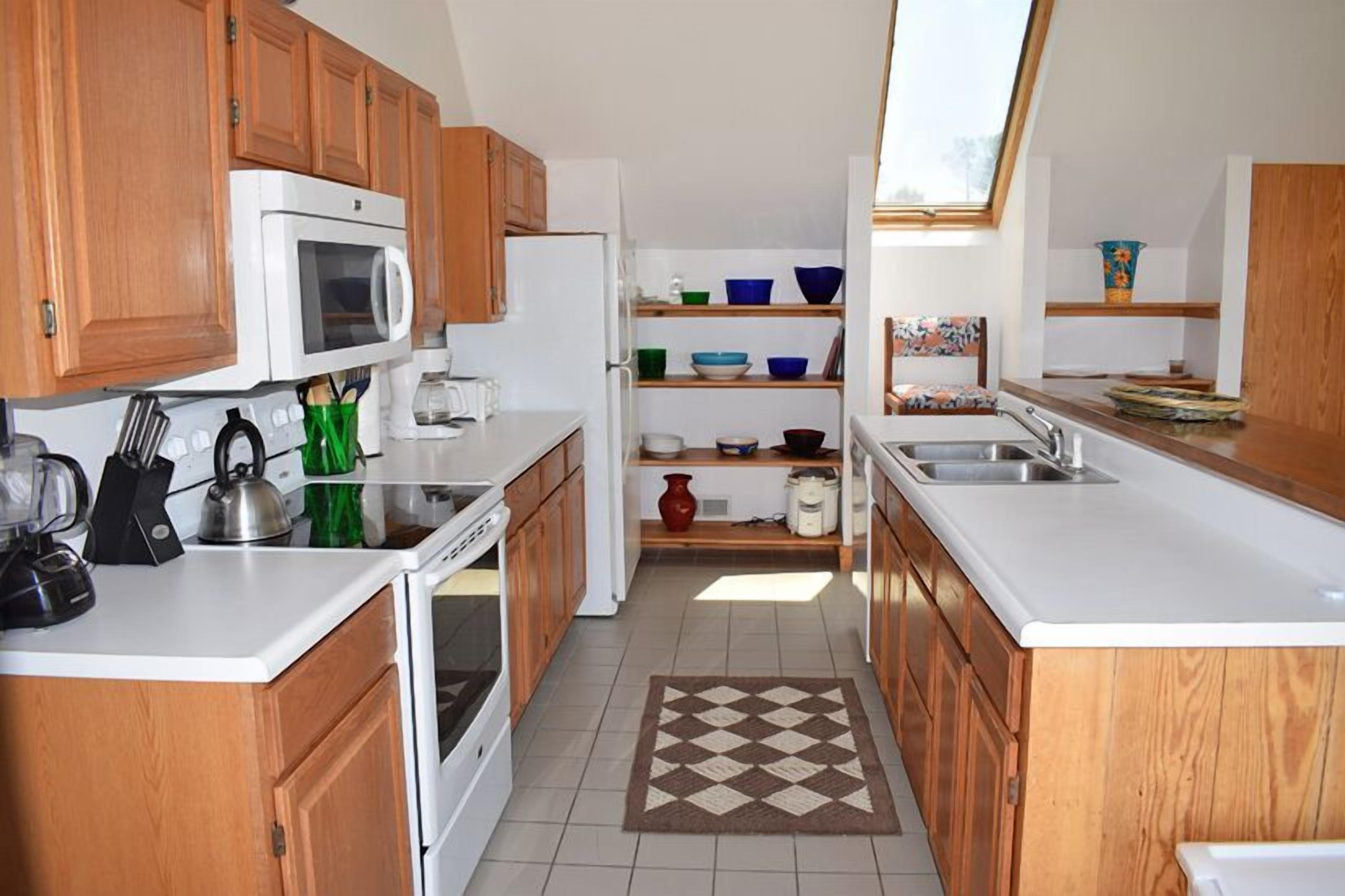 A kitchen with wooden cabinets, white countertops, a microwave, a kettle, and a variety of bowls and jars on shelves.