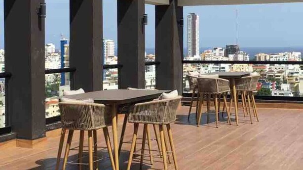 Una terraza en la azotea con suelo de madera, mesas y sillas de mimbre y vistas al horizonte de la ciudad.
