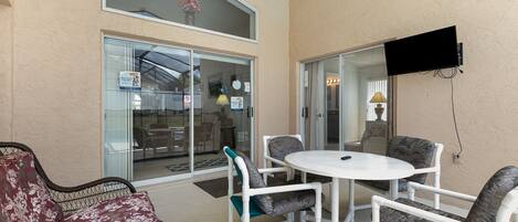 A patio area with a white table and four chairs, a wicker sofa, and a TV mounted on the wall.