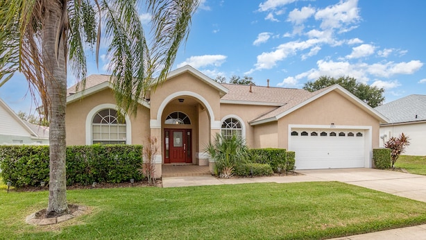 A single-story house with a red door, a garage, and a well-maintained lawn. - Orange Tree Pool Home with spa. Enjoy recreation and relaxation in this inviting community near the best of Orlando's hot spots! (Clermont)