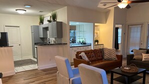 Uma sala de estar moderna com um sofá de couro marrom, duas poltronas e uma mesa de centro. Cozinha com armários brancos e uma área de jantar com mesa e cadeiras são visíveis ao fundo.