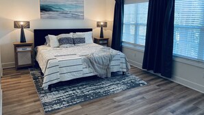 Quarto com cama, duas mesinhas de cabeceira, ventilador de teto, janela com cortinas e um quadro na parede.