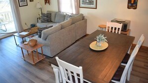 A cozy living room with a wooden dining table, white chairs, a grey sofa, and a small side table with decorative items. - Pony Swim Ridge- Waterviews, Close to Memorial Park (Chincoteague)