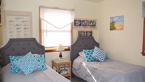 A bedroom with two single beds, a nightstand with a lamp, a framed shark poster, and a beach painting. - Pony Swim Ridge- Waterviews, Close to Memorial Park (Chincoteague)