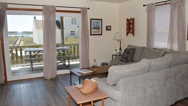 A living room with a grey sofa, a wooden coffee table, a lamp, and a view of a dock and water outside. - Pony Swim Ridge- Waterviews, Close to Memorial Park (Chincoteague)