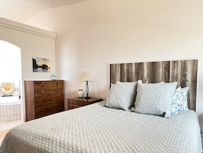 Une chambre à coucher avec un grand lit, une tête de lit en bois, une table de chevet avec une lampe, une commode et une étagère murale agrémentée d’objets décoratifs.
