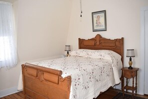 A bedroom with a wooden bed, floral bedspread, two bedside lamps, a framed picture on the wall, and a window with a sheer curtain.