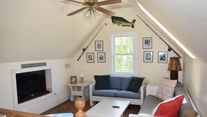 A cozy attic bedroom with a sloped ceiling, a ceiling fan, a large window, a sofa, a TV, and a wooden staircase. - Sailer Moon- 4 BR | Walk to Town | Sunset Views (Chincoteague)