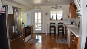 A kitchen with stainless steel appliances, wooden cabinets, and a window with curtains. - Sailer Moon- 4 BR | Walk to Town | Sunset Views (Chincoteague)