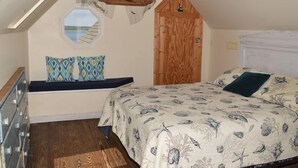 A bedroom with a sloped ceiling, a wooden door, a bed with a floral bedspread, and a window with a view of the sea. - Sailer Moon- 4 BR | Walk to Town | Sunset Views (Chincoteague)