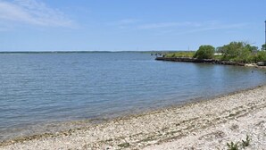 A pebbly beach with calm water, a distant pier, and sparse vegetation. - Sailer Moon- 4 BR | Walk to Town | Sunset Views (Chincoteague)