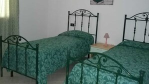 Una stanza con due letti in metallo, una coperta a letto fantasia verde, una parete bianca, un quadro con un’ancora, una lampada a sospensione e una finestra con tenda verde.