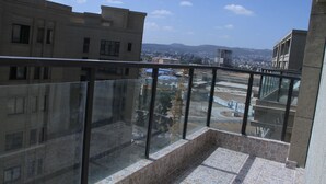 Un balcone con pavimento in pietra e ringhiera in vetro, affaccia su un panorama urbano con edifici e una catena montuosa in lontananza.