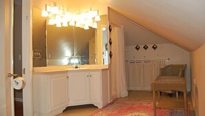 Ein Badezimmer mit einem großen Spiegel, weißen Schränken, einer Toilette und einem Korbstuhl.