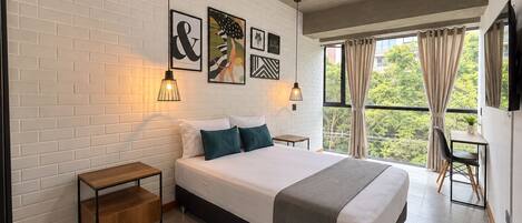 A modern bedroom with a large bed, bedside tables, artwork on the wall, and a view of greenery through the window.