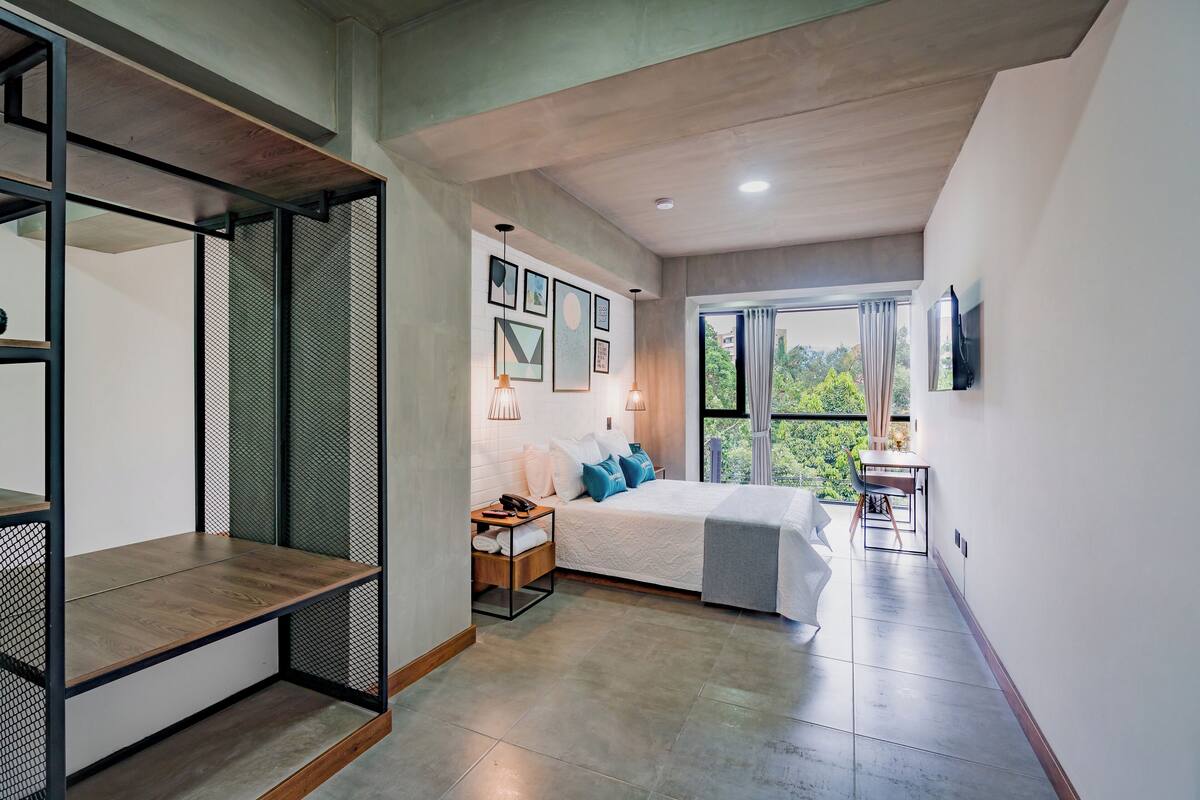 A modern bedroom with a bed, bedside table, and a view of greenery.