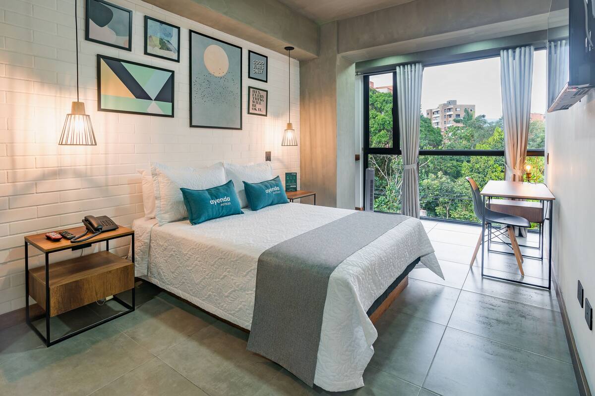A modern bedroom with a bed, bedside tables, a desk, and a view of greenery through the window.