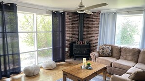 A living room with a brick fireplace, a ceiling fan, a wooden coffee table, a brown sofa, and a view of greenery outside. - West Avenue Place, quiet neighborhood, close to downtown  (Cody)