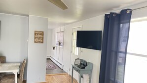 A room with a wooden floor, a dining table with chairs, a TV, and a small table with a sign. - West Avenue Place, quiet neighborhood, close to downtown  (Cody)
